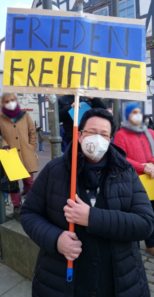 Frau mit FFP2-Maske hält Plakat an einer Stange. Das Plakat zeigt die ukrainische Flagge (oben blau unten gelb). Darauf steht geschrieben: Frieden Freiheit.