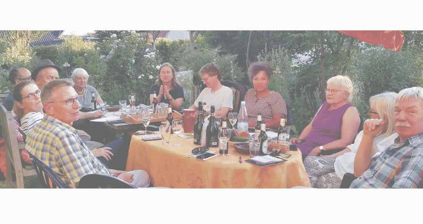 Gruppenfoto der Ortsgruppe. Die Mitglieder sitzen an einem Sommertag am Tisch. Auf dem Tisch stehen u.a. Schreibsachen, Handys, Gläser und Trinkflaschen.